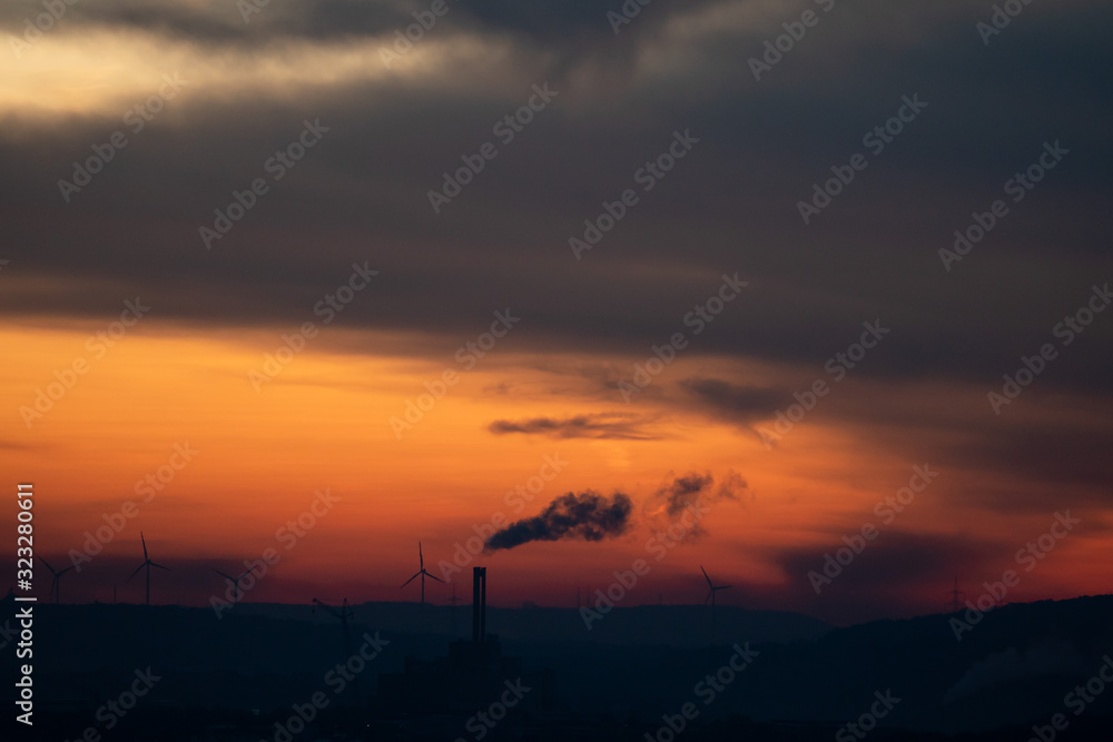 The silhouette of a smoking chimney in the far distance in front of a dramatic red sunrise and some wind turbines