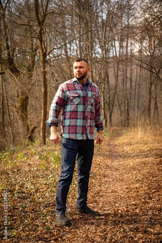 Bearded hipster man with serious face is thinking about something while standing outdoors against autumn fall with copy space, stylish brutal male with trendy look walking in park at sunset. © Inception