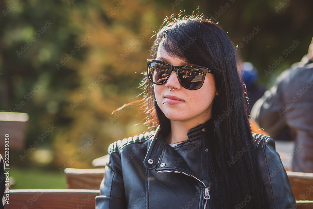Cute sexy young woman with long black hair, sunglasses, septum piercing and a leather jacket sitting on a bench in a park and enoying the sun. Fetish style woman in leather outdoor Cute sexy young woman with long black hair, sunglasses, septum piercing and a leather jacket sitting on a bench in a park and enoying the sun. Fetish style woman in leather outdoor