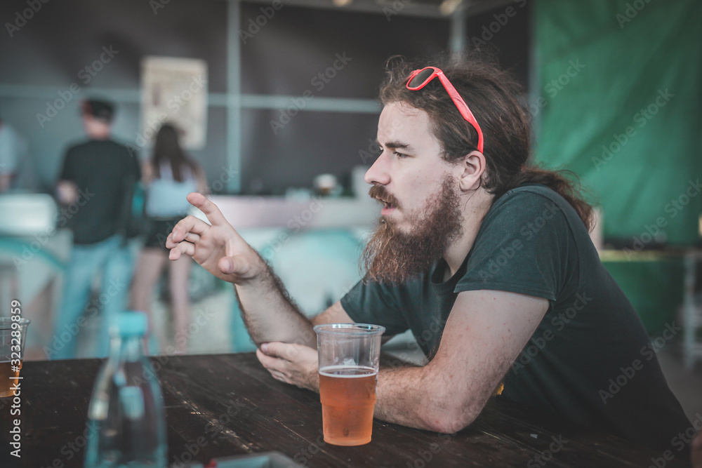 A hipster looking man with red sunglasses holding a fictive object while sitting at a table with some beers on an event.