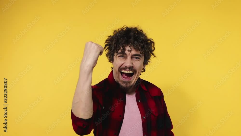 Man celebrates team victory. Closeup portrait of happy smiling guy with curly hair looking at camera isolated on yellow background. Slow motion. Man celebrates team victory. Closeup portrait of happy smiling guy with curly hair looking at camera isolated on yellow background. Slow motion.