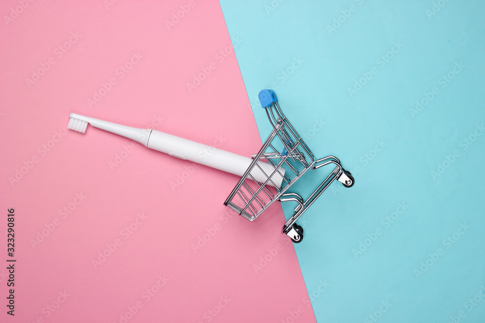 Mini shopping trolley with electric toothbrush on a pink-blue pastel background. Top view.