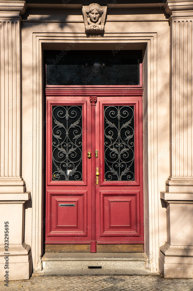 Classical European doorway with stone pillars around red door with gold details,. Ornate details in dark windows and stone figurine over entrance. Shot in bright daylight