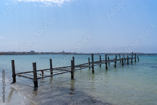 The beautiful Italian island of Sardinia.  The beach and shoreline before the historic city of Alghero.  A metal jetty leading out to sea. 