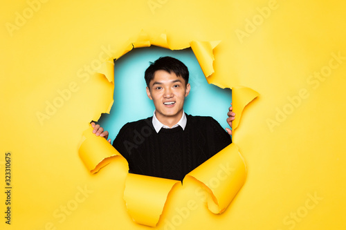 Young chinese man keeps head in hole of torn yellow wall. Male head in ripped paper. © dianagrytsku