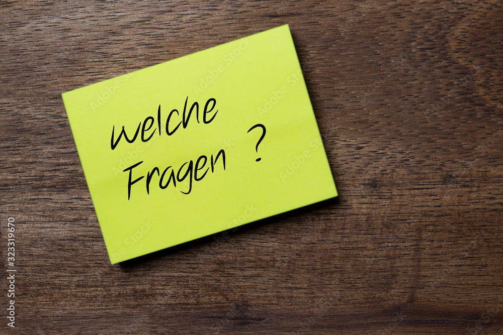 The question in german: Which questions? You can find on a yellow self-adhesive note sticker diagonally on a dark wooden table top with stylized handwriting. The question in german: Which questions? You can find on a yellow self-adhesive note sticker diagonally on a dark wooden table top with stylized handwriting.