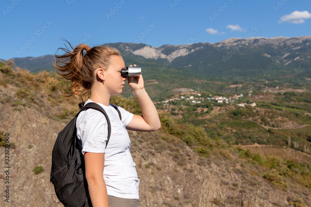 Girl hiker looking in binoculars enjoying spectacular view on mountains and blue sky background. Summer landscape. Adventure or travel alone with backpack. Hipster lifestyle. Young traveler in nature