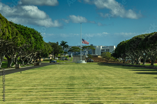 Oahu, Hawaii, USA. - January 10, 2012: Wide shot over green National Memorial Cemetery of the Pacific with flag half pole under blue cloudscape. Green trees on side, and white building in back.