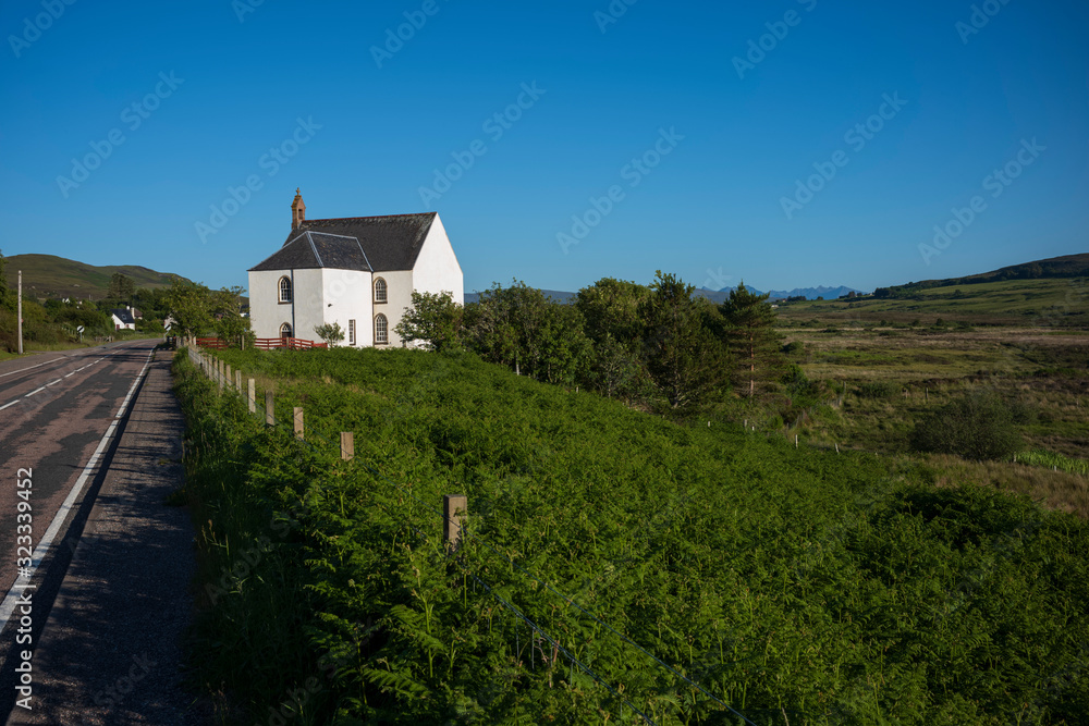 A small white church along the side of a country road in the Isle of Skye, Scotland, is illuminated by the daylight on a cloudless, summer day.