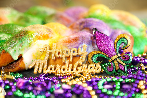 Fotografiet Happy Mardi Gras text in gold glitter and a king cake with yellow, green, and purple sprinkles surrounded by Mardi Gras beads and a glittering fleur de lis