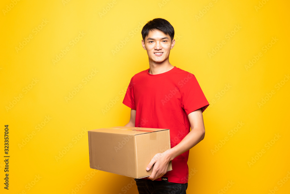 Portrait of handsome chinese deliver holding card-board box, showing thumbs-up, isolated over yellow background Portrait of handsome chinese deliver holding card-board box, showing thumbs-up, isolated over yellow background