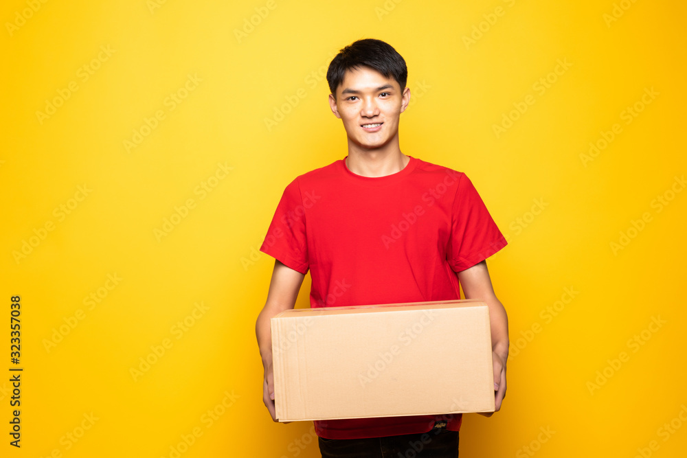 Portrait of handsome chinese deliver holding card-board box, showing thumbs-up, isolated over yellow background Portrait of handsome chinese deliver holding card-board box, showing thumbs-up, isolated over yellow background