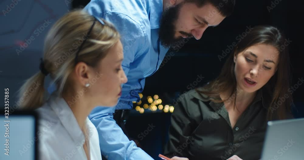 Caucasian male boss checking work of two women office workers and explaining his ideas. Man teaching young female co-workers business and showing them strategy late in evening. Teamwork at company. Caucasian male boss checking work of two women office workers and explaining his ideas. Man teaching young female co-workers business and showing them strategy late in evening. Teamwork at company.