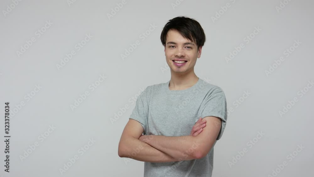Happy handsome brunette man in T-shirt turning to camera and putting hands crossed, looking with toothy smile, expressing optimism, positive outlook. indoor studio shot isolated on gray background Happy handsome brunette man in T-shirt turning to camera and putting hands crossed, looking with toothy smile, expressing optimism, positive outlook. indoor studio shot isolated on gray background