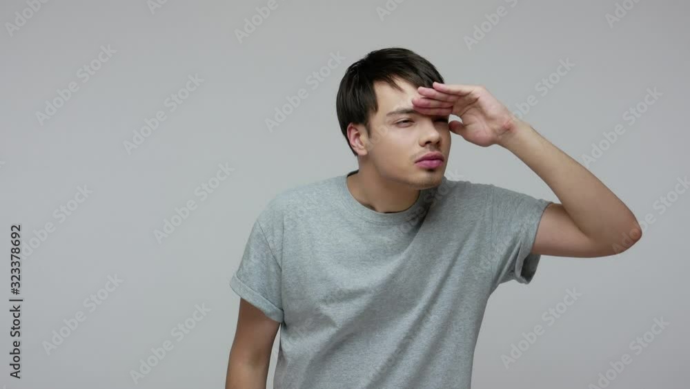 Young brunette man in T-shirt looking around with attentive concentrated face, holding palm over eyes and viewing distance, watching forward future. indoor studio shot isolated on gray background