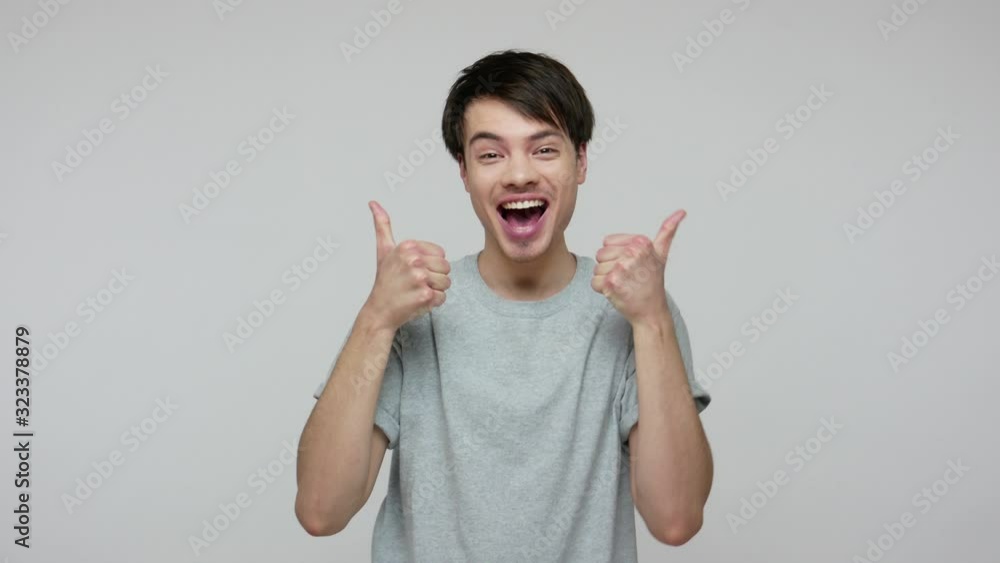Excited young brunette man in T-shirt shouting Yeah, enjoying success and showing thumbs up, double like gesture, satisfied with awesome service. indoor studio shot isolated on gray background Excited young brunette man in T-shirt shouting Yeah, enjoying success and showing thumbs up, double like gesture, satisfied with awesome service. indoor studio shot isolated on gray background