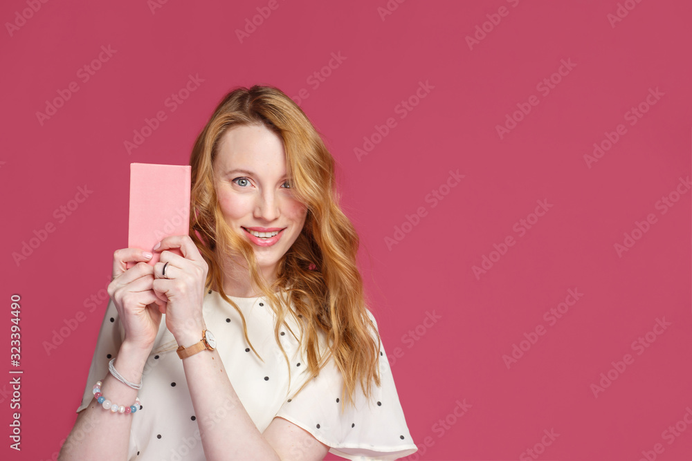 Cheerful blonde girl holds a pink notebook in her hands, looks at the camera and smiles, isolated on a pink background with copy space. Cheerful blonde girl holds a pink notebook in her hands, looks at the camera and smiles, isolated on a pink background with copy space.