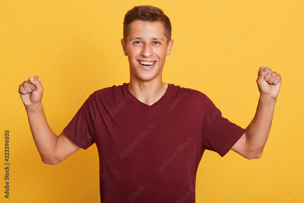 Indoor shot of cheerful handsome attractive guy raising arms, clenching fists, making gesture, having sincere facial expression, wearing dark red t shirt, standing isolated over yellow background. Indoor shot of cheerful handsome attractive guy raising arms, clenching fists, making gesture, having sincere facial expression, wearing dark red t shirt, standing isolated over yellow background.