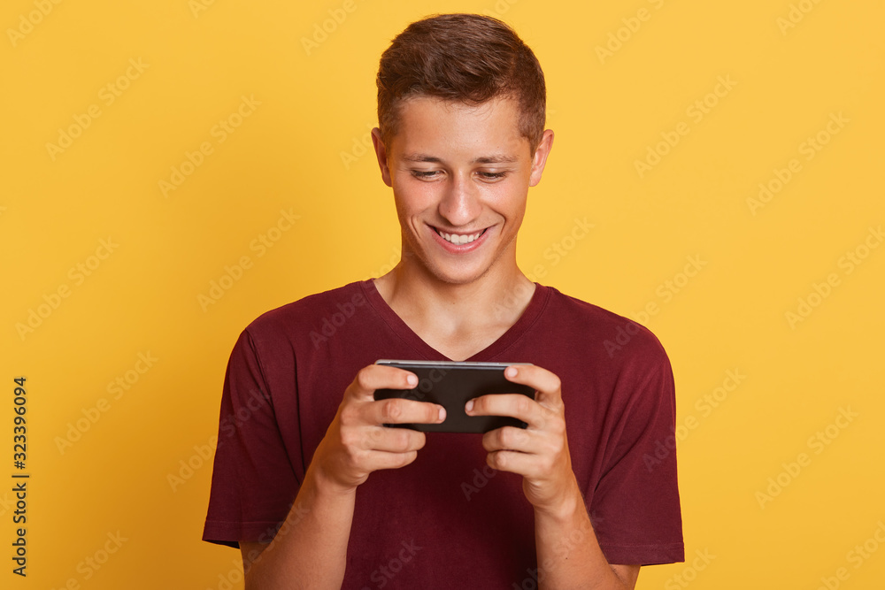 Image of cheerful sincere good looking guy playing games, using his smartphone, looking attentively at screen, having pleasant facial expression, wearing dark red t shirt. Technologies concept. Image of cheerful sincere good looking guy playing games, using his smartphone, looking attentively at screen, having pleasant facial expression, wearing dark red t shirt. Technologies concept.