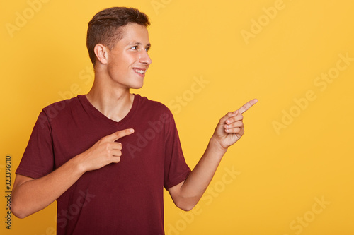 Image of delighted inspired young guy looking aside, making gesture, raising his arms, wearing red t shirt, being in good mood, showing direction with his forefingers. Copyspace for advertisement.
