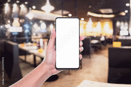 Mockup image of hand holding white screen mobile phone for your advertisement with blurred background of restaurant. Mock up modern smartphone in a shop.