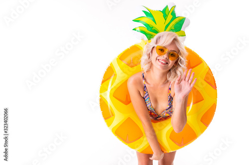 Fototapeta Naklejka Na Ścianę i Meble -  Pretty blonde caucasian female stands in swimsuit with rubber beach pineapple ring and smiles isolated on white background