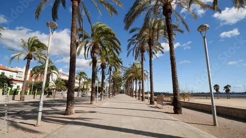 Beach seaside promenade surrounded with tall palm trees moving because of wind on hot sunny summer day, with blue sky and clouds. Concept of holidays and vacation. Spain, Vilanova i la Geltru photo