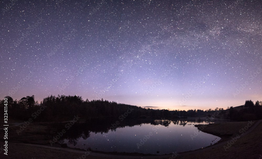 Milky Way over a lake with stars reflecting in the water with a purple sky and surrounded by trees