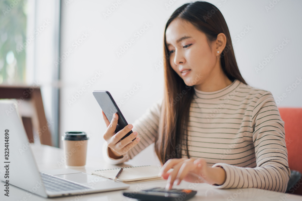 Young woman using a calculator and other hand using phone to chat with customer and have notebook on the table at cafe or co working space which smiling and felling happy. co working space concept. Young woman using a calculator and other hand using phone to chat with customer and have notebook on the table at cafe or co working space which smiling and felling happy. co working space concept.