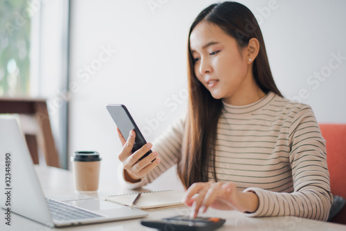 Young woman using a calculator and other hand using phone to chat with customer and have notebook on the table at cafe or co working space which smiling and felling happy. co working space concept.