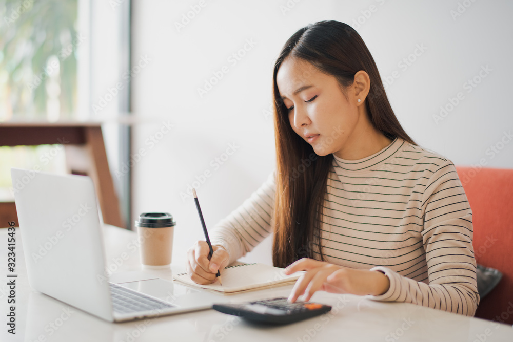 Young Asian woman using a calculator and other hand using pencil take a note in the book and have notebook on the table at cafe or co working space. co working space concept. Young Asian woman using a calculator and other hand using pencil take a note in the book and have notebook on the table at cafe or co working space. co working space concept.