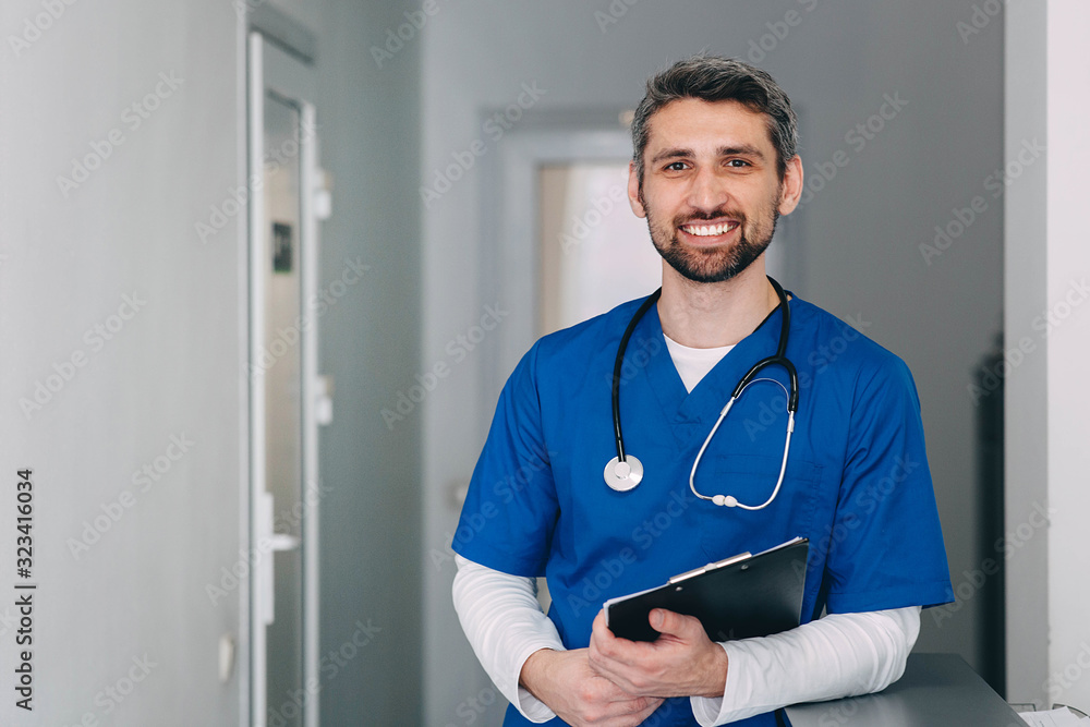 male nurse with stethoscope standing at clinic. He is smiling and looking at the camera. - obrazy, fototapety, plakaty 