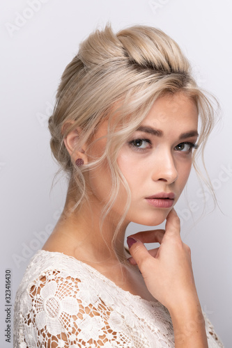 A large head portrait of a beautiful young girl with dyed blonde hair arranged in a hairstyle, with makeup and beautiful lips and teeth. Hands near the face. a neutral gray background