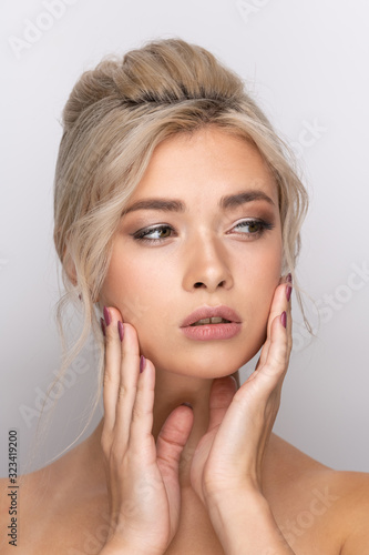 A large head portrait of a beautiful young girl with dyed blonde hair arranged in a hairstyle  with makeup and beautiful lips and teeth. Hands near the face. a neutral gray background