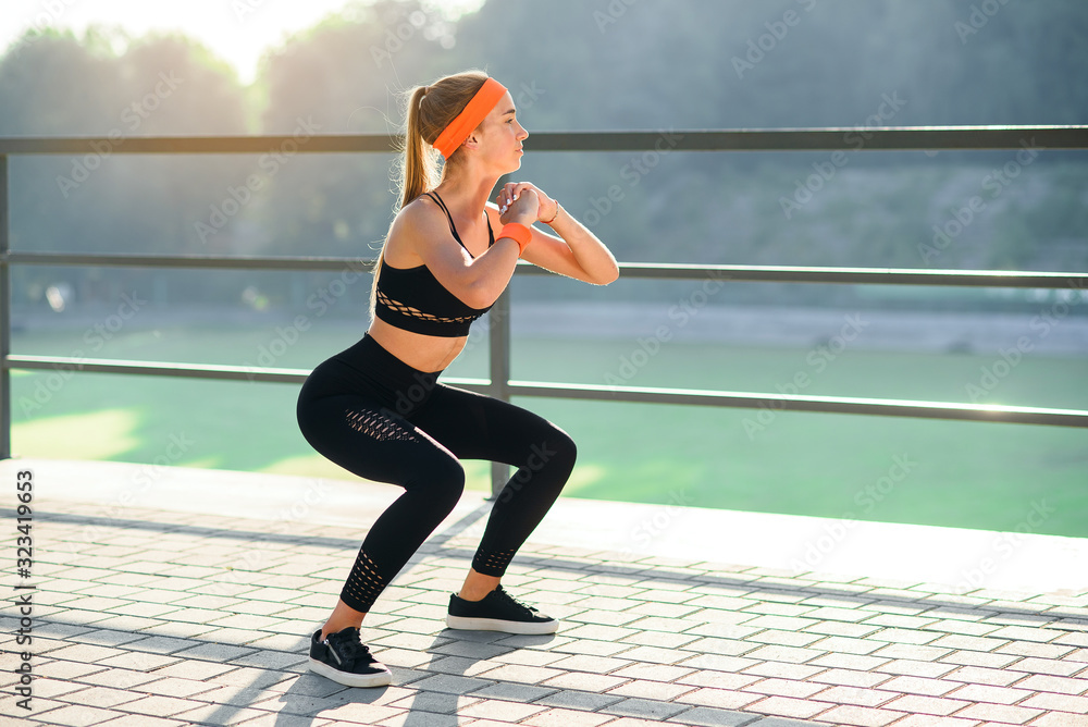 Gorgeous sporty girl in black sportswear and orange headband which squating  during fitness training at stadium outdoors. Stock Photo | Adobe Stock