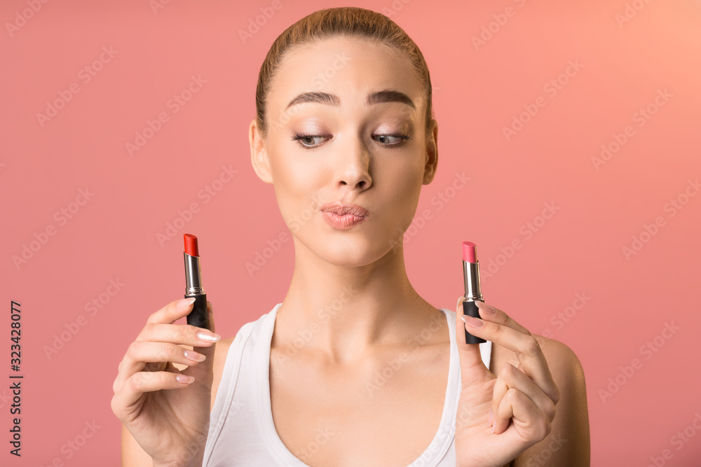 Fototapeta premium Beautiful Woman Choosing Lipstick For Makeup Standing On Pink Background