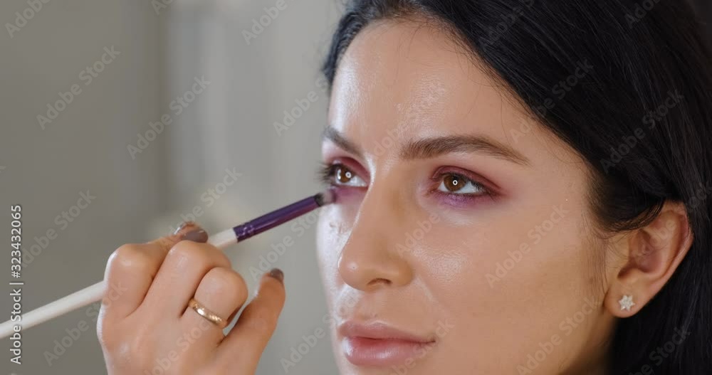 Portrait of brunette girl make-up artist applies heavy shadows with brush from palette to eyelids in makeup studio, against background of bokeh light bulbs on mirror. Beauty lamp. Makeup and beauty Portrait of brunette girl make-up artist applies heavy shadows with brush from palette to eyelids in makeup studio, against background of bokeh light bulbs on mirror. Beauty lamp. Makeup and beauty