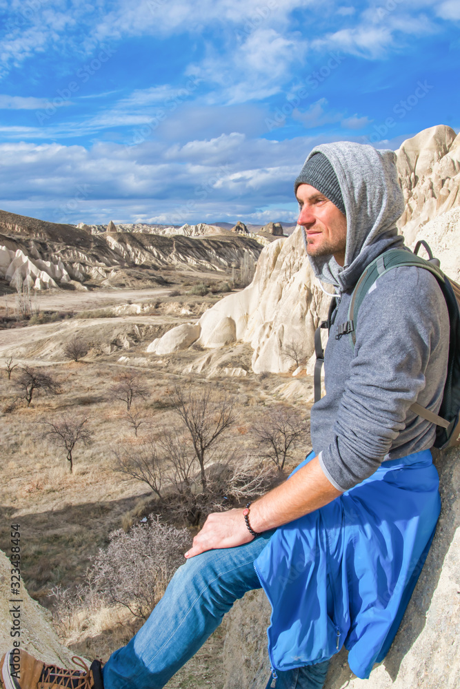Naklejka premium Male tourist sits and poses for a photo in red valley in Kapadokya with rocky formations in the background. Exploration of interesting geologcal sites.
