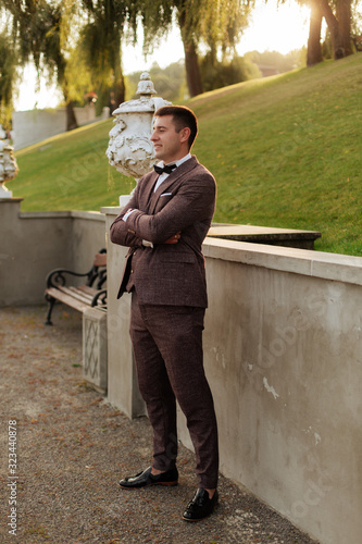 Lifestyle portrait of happy groom posing for camera outdoor at nature with oak tree on background. Cheerful fiance with smiling face in jakcet, bowtie and white shirt portrait before wedding ceremony photo