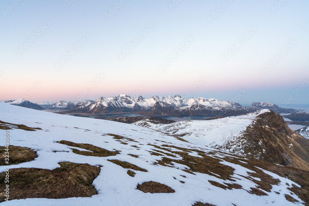 Panorama of Landscape shot from Ryten towards fredvang and ramberg in Lofoten island in Norway during blue hour. Snow cover the peaks and city lights in the background.