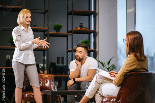 young caucasian couple, blond lady and man discuss their relationship and problems with professional psychologist, woman stand talking animatedly and gesturing, man sit and listen