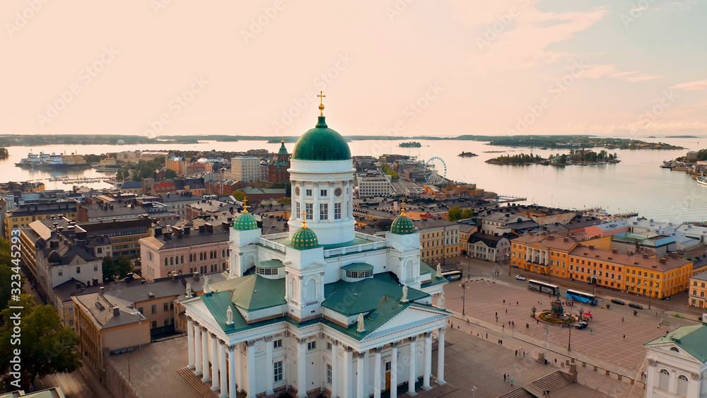 A view of Helsinki from a height above a white church with green domes, from which the whole city, its houses, small islands, a ferris wheel and a bay are clearly visible