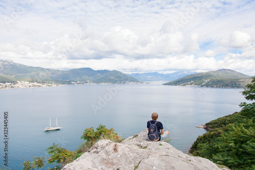 Traveler with backpack sitting on mountain top above sea beach. Tourist enjoying traveling, summer vacation, adventure, relaxation, freedom. Man has rest. Copy space. Rear view.