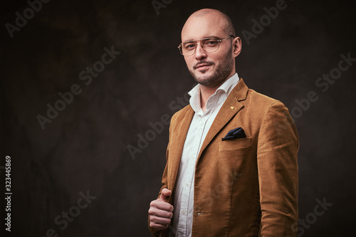 Successfull well-dressed mature bald businessman posing for camera in a dark studio wearing stylish mustard color velvet jacket, white shirt and glasses
