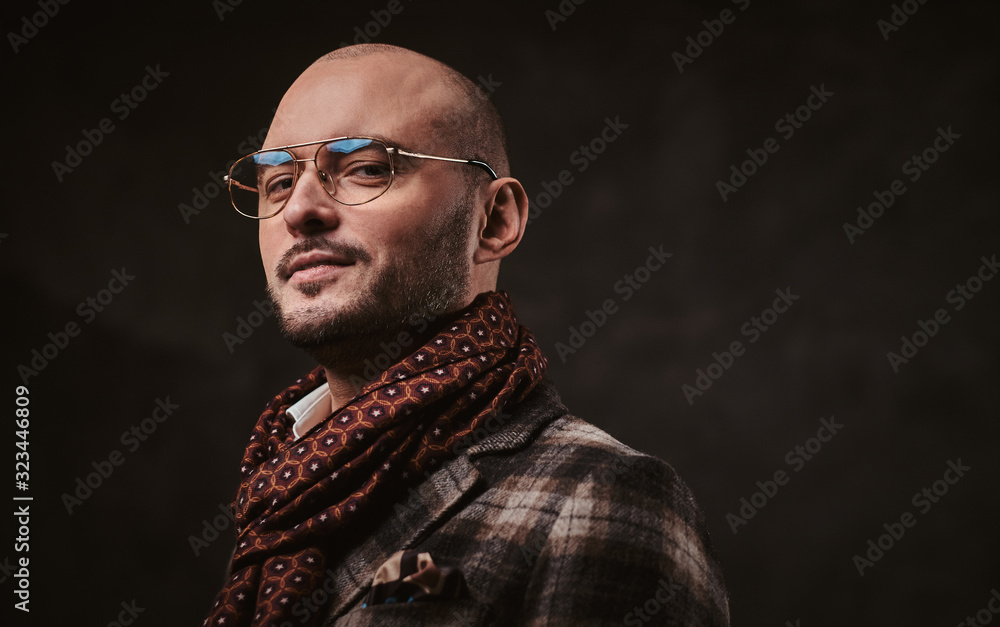 Fototapeta premium Smirking and confident stylish bald businessman posing for camera in a dark studio wearing checkered wool jacket, glasses and ruby patterned scarf