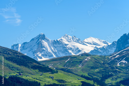 View of beautiful landscape in the Alps with fresh green meadows and snow-capped mountain tops in the background on a sunny day with blue sky and clouds in springtime.