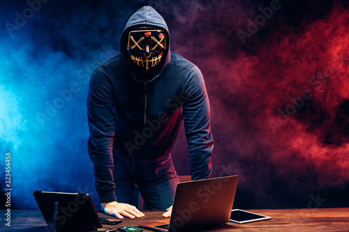 portrait of an anonymous man hiding his face behind a scary neon mask in a colored smoke. male in pullover stand in scary mask isolated over dark smoky space. modern devices, laptop on table. photo