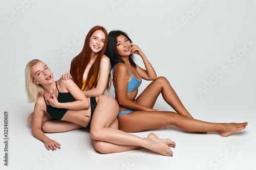 Young women with slim bodies in swimsuits posing to the camera while sitting on the floor isolated on white, free time, spare time, lifestyle, copy space © the faces