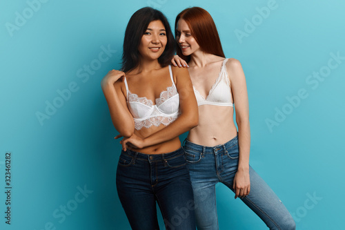 smiling ginger girl leaning on her friend and wispering something to her , close up photo, isolated blue background, studio shot, women sharing secrets photo