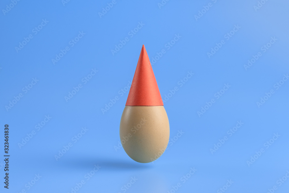 Obraz premium One brown egg in a red festive cap on a blue background. Minimal Happy Easter concept decoration. Copy space for text mockup